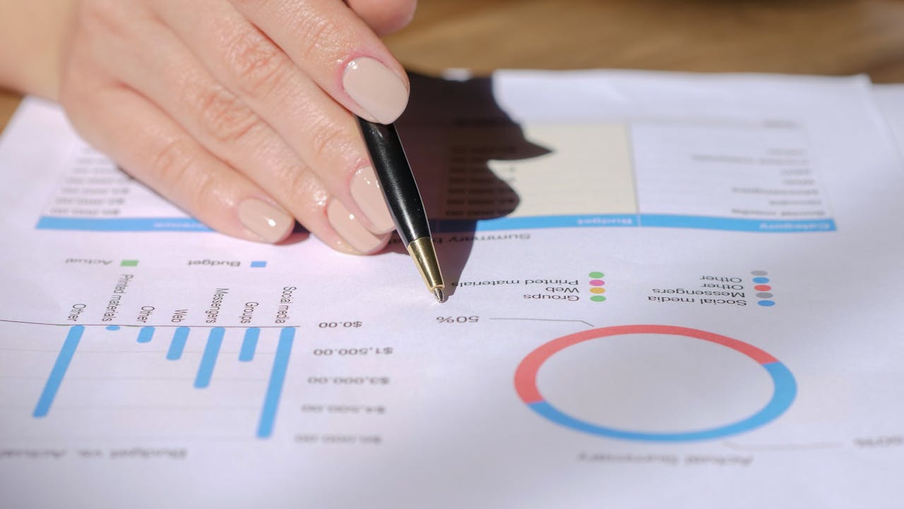 Close-up of a hand with a pen pointing at a financial document with graphs and charts.